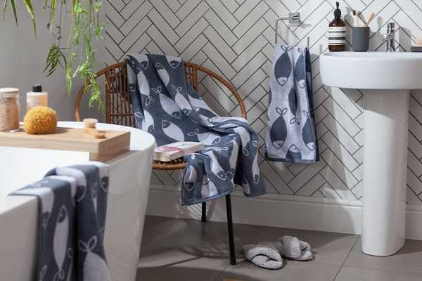 A white herringbone tiled bathroom with two-tone fish print towels laid out on a bamboo chair.